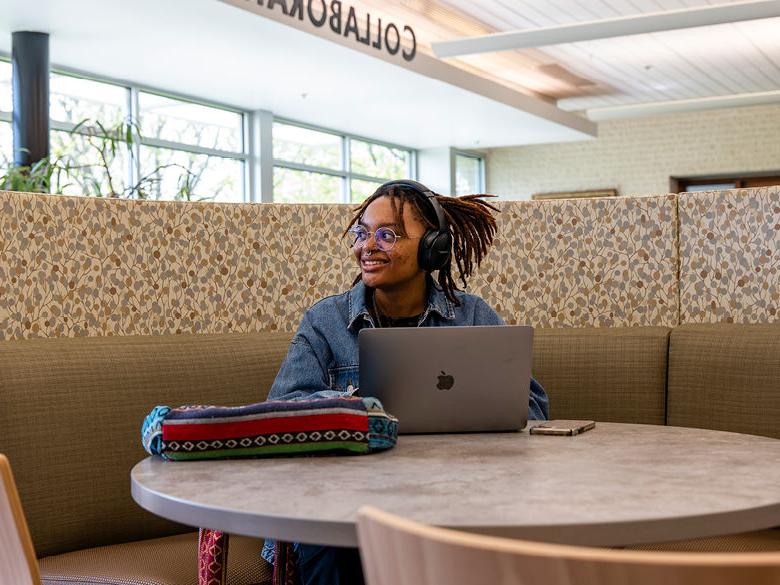 Smiling female student sits with Mac laptop at table in library.